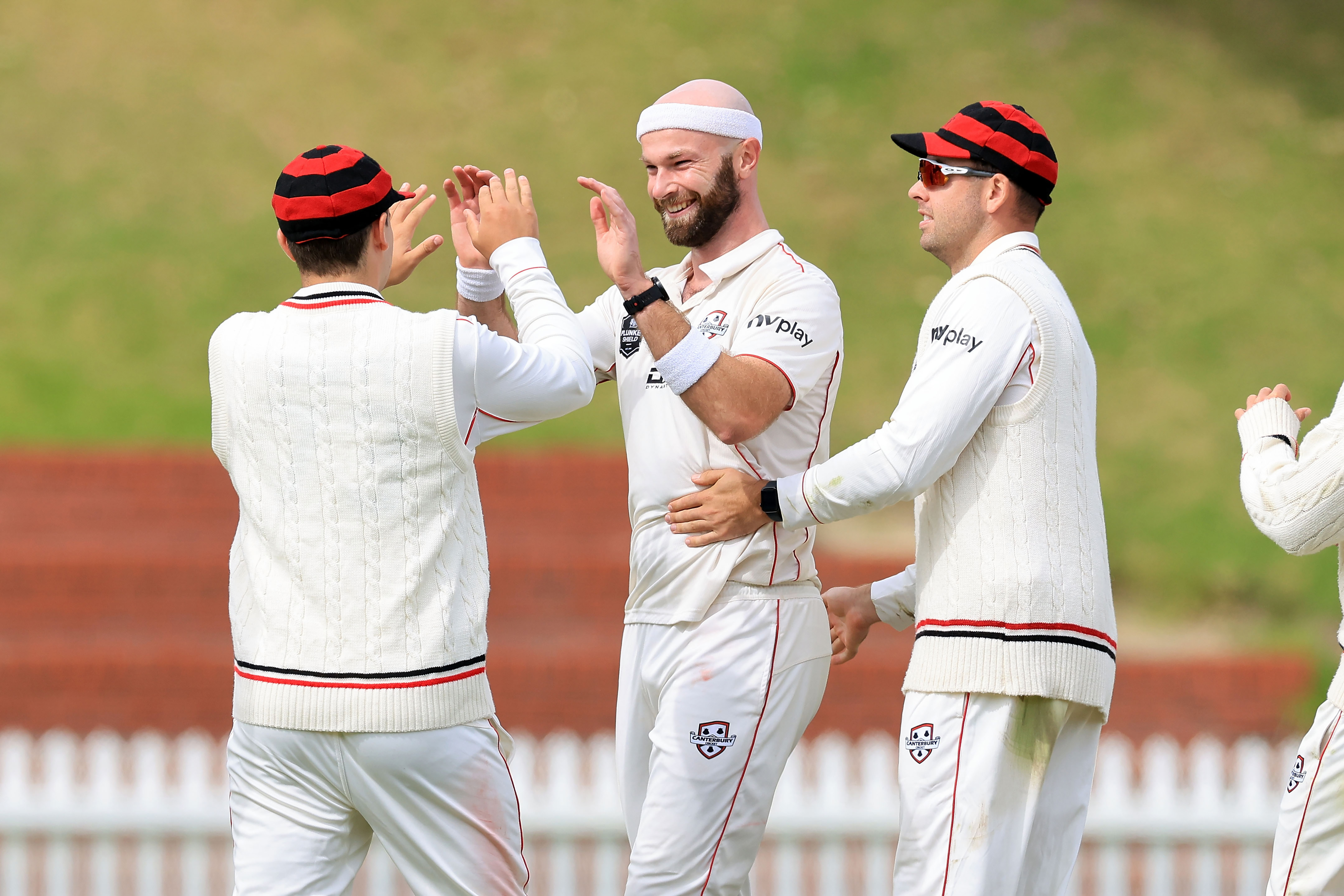 Three Canterbury Cricket players, two high-fiving after a wicket, NV Play logo on sleeves.